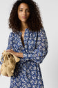 Woman with curly hair wears a blue and white floral dress, holding a beige woven bag with tassels, standing against a plain light background.