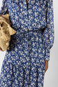 A person in a blue floral dress with long sleeves, holding a woven straw bag with tassels. Only the upper body and dress details are visible.