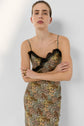 Person with light skin and brown hair pulled back, wearing a sleeveless robe longue à motif floral with thin straps and black dentelle trim, stands with arms crossed and a neutral expression against a plain light background.