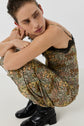 A person in a robe longue à motif floral avec une délicate dentelle noire crouches, looking to the side. They wear black heeled boots and gold rings, with hair pulled back. The background is plain and light-colored.