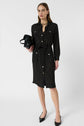 A woman wearing a black, long-sleeve, button-down robe chemise à pois crafted in soft viscose haboutaï, black pointed shoes, and holding a black handbag stands against a plain white background.