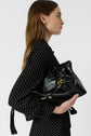 Woman with long brown hair in a robe chemise à pois noire—a stylish black dress with white polka dots in soft viscose haboutaï—holding a large black leather handbag with gold buckles, standing in profile against a plain light background.