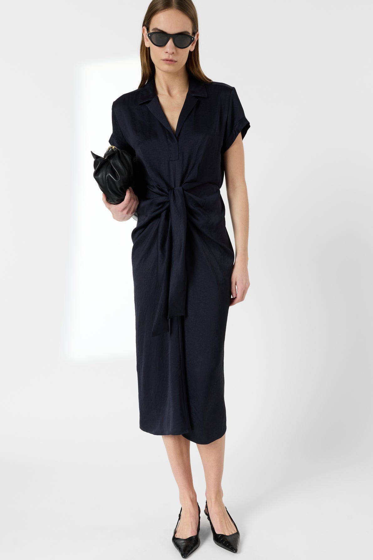 A woman wearing sunglasses, a short-sleeve navy dress with a front tie, and black pointed-toe heels holds a black clutch. She stands against a plain white background.