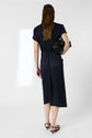 A person with long brown hair, wearing a navy midi dress and black heels, stands facing away while holding a black handbag under their right arm against a plain white background.