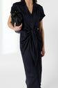 A person wearing a short-sleeve, navy blue, collared midi dress with a front tie detail, holding a black clutch bag under their arm, standing against a plain white background.