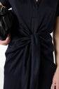 A person in a dark, textured dress with a front knot detail holds a black clutch bag. Only the midsection and part of one arm are visible against a plain background.