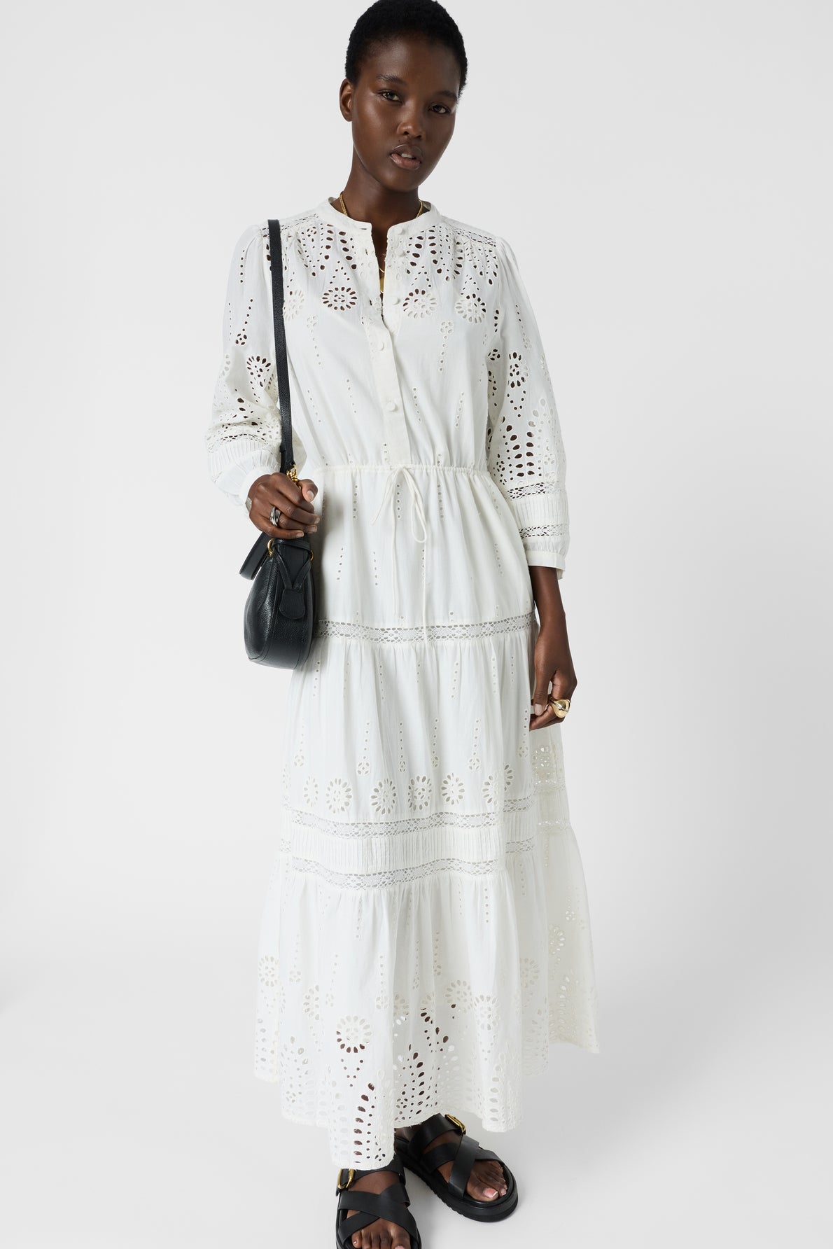 A person wearing a long white eyelet dress, black sandals, and a black crossbody bag stands against a plain white background.
