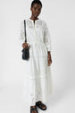 A person wearing a long white eyelet dress, black sandals, and a black crossbody bag stands against a plain white background.