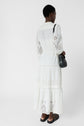 Person standing with their back to the camera, wearing a long white eyelet dress, black sandals, and carrying a small black shoulder bag. The background is plain white.