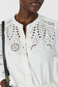 A person wearing a white eyelet-pattern blouse with round buttons, gold necklace, and carrying a black purse. The blouse features intricate cutout designs on the chest and shoulders.