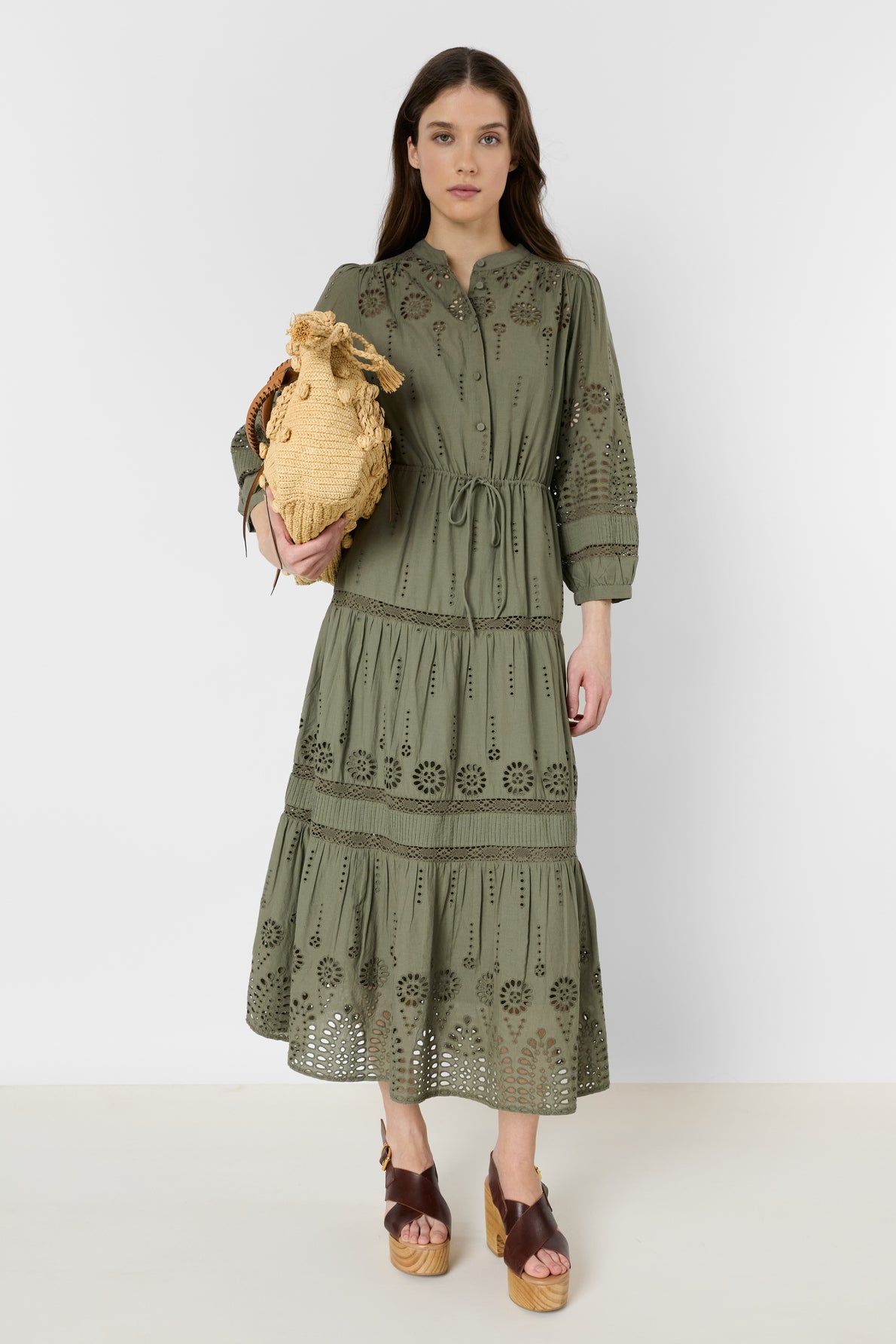 Woman wearing a robe longue en coton kaki à broderies ajourées, holding a woven straw bag and wearing brown platform sandals, standing against a plain white background.