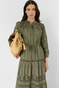 Woman wearing a coton kaki robe longue with broderies ajourées and long sleeves, holding a beige woven bag with tassels against a plain background.