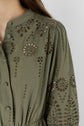 Close-up of a person wearing an olive green blouse in coton kaki with embroidered floral cutout patterns, reminiscent of broderies ajourées, and fabric-covered buttons along the front. The sleeve and shoulder details are visible.