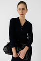 A woman with tied-back hair in a ribbed navy robe longue à col polo holds a large black leather bag close to her chest while looking seriously at the camera against a plain white background.