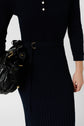 Person wearing a navy blue, long-sleeve maille côtelée dress with a belt and three buttons on the chest, featuring a chic col polo neckline. They are holding a black leather handbag with gold accents. Only the upper body is visible.