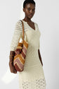 A woman in a cream crochet dress stands holding a woven bag with zigzag patterns in brown, gold, and pink, featuring pom-pom accents and a brown strap. She wears gold bangles and looks at the camera.
