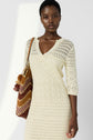 A woman wearing a cream-colored, crocheted dress with three-quarter sleeves holds a brown and yellow zigzag-patterned bag over her shoulder, standing against a light background.