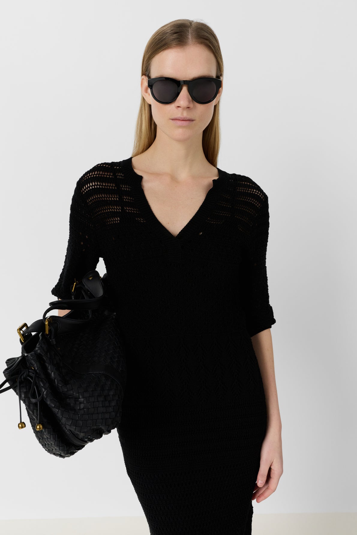 A woman wearing a black knit dress and large black sunglasses holds a textured black handbag. She has straight, light brown hair and stands against a plain white background.