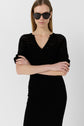 Woman wearing a black knit dress and black sunglasses stands with arms crossed against a plain white background.