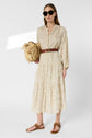 Woman wearing sunglasses, a robe longue beige à motifs avec une ceinture marron, des sandales brunes et tenant un sac en paille tressée, adopte un style bohème tout en se tenant contre un fond blanc uni.