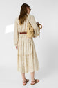 A woman in a cream robe longue with long sleeves and a brown belt stands facing away, carrying a woven straw bag and wearing sandals. Her hair is loose and she exudes a style bohème vibe in the brightly lit studio setting.