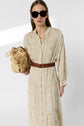 Woman wearing a cream robe longue with fleurs brodées and a brown belt, holding a woven bag. She has long hair, is wearing large dark sunglasses, and stands against a plain white background, effortlessly capturing the essence of style bohème.