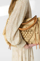 A person in a long-sleeved, cream-patterned robe longue adorned with fleurs brodées holds a woven straw tote bag with leather handles and textured pom-pom details, embodying un style bohème. The background is plain and bright.