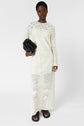 A person in a long-sleeve, white lace dress stands against a white background, holding a black clutch bag and wearing black sandals.