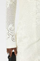 A person wearing a white, long-sleeved, lace-patterned dress holds a small black handbag. The dress features floral and circular cut-out designs. Only the arm, hand, and part of the dress are visible.