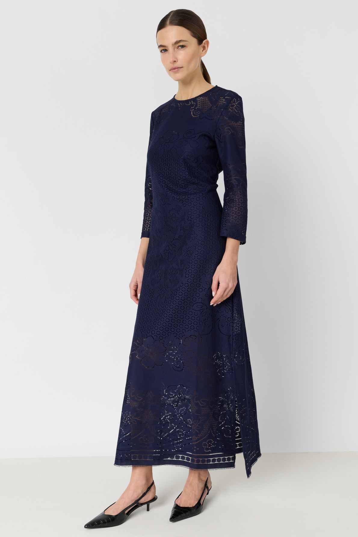 Woman wearing a long-sleeved, navy blue lace dress with a fitted bodice and flared skirt—an exquisite robe longue dentelle in a rich robe bleu encre—standing against a plain white background and wearing black pointed-toe heels.