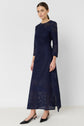 Woman wearing a long-sleeved, navy blue lace dress with a fitted bodice and flared skirt—an exquisite robe longue dentelle in a rich robe bleu encre—standing against a plain white background and wearing black pointed-toe heels.