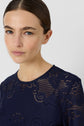 A woman with straight brown hair pulled back, wearing a robe bleu encre en dentelle, looks at the camera with a neutral expression against a plain light background.