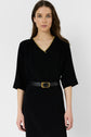 Woman with long brown hair wearing a black dress with elbow-length sleeves, a V-neck with yellow trim, and a black belt with a gold buckle, standing against a plain white background.