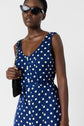 Woman wearing cat-eye sunglasses and a sleeveless blue dress with white polka dots, holding a black purse with a gold chain over her shoulder, standing against a plain white background.