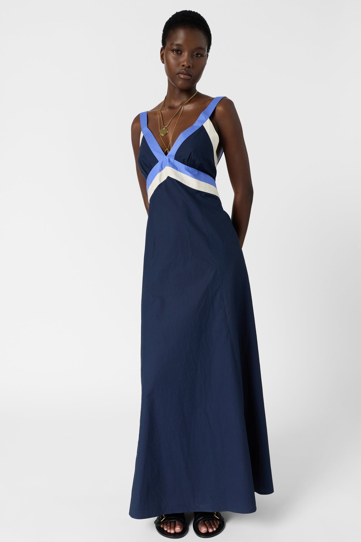 Woman in a navy blue robe longue, reminiscent of the élégance des années 50, featuring white and light blue chevron stripes at the neckline. Crafted in coton tricolore, she pairs the look with black sandals and layered gold necklaces, standing against a plain white background.