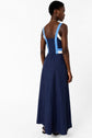 A woman stands facing away, wearing a robe longue inspired by the années 50, featuring a sleeveless navy maxi silhouette with light blue straps and panel details. The dress has a fitted bodice and flows to the floor. She has short hair and is wearing a necklace.
