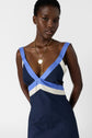 A woman wearing a sleeveless navy robe longue with blue and white accents reminiscent of coton tricolore and layered gold necklaces stands against a plain white background.