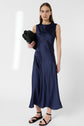 Woman wearing a sleeveless, navy blue satin midi dress with black sandals, holding a black clutch under her arm, standing against a plain white background.