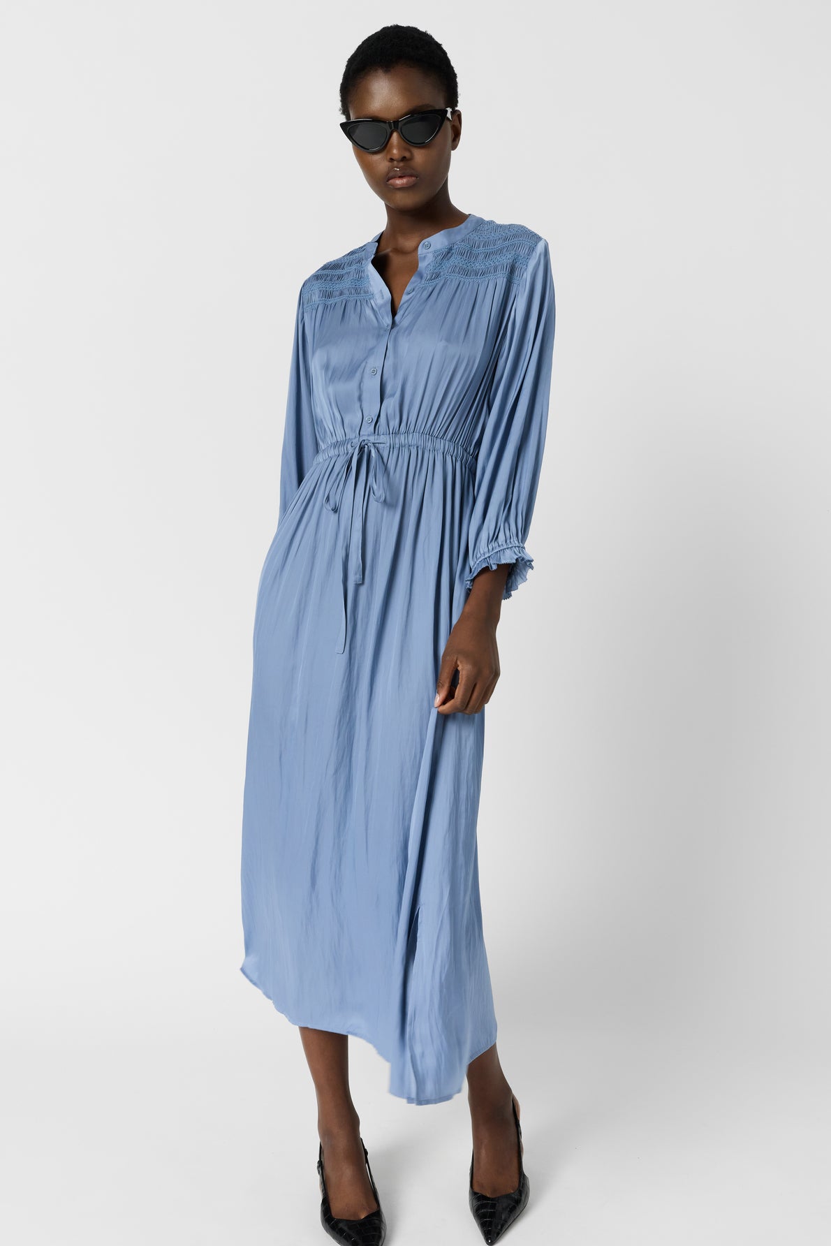 Woman wearing a long-sleeve, light blue midi dress with a cinched waist, black pointed heels, and dark sunglasses standing against a plain white background.
