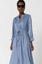 A woman wearing a light blue, long-sleeved, belted midi dress with a gathered waist and lace detail on the shoulders, accessorized with black cat-eye sunglasses and gold hoop earrings.