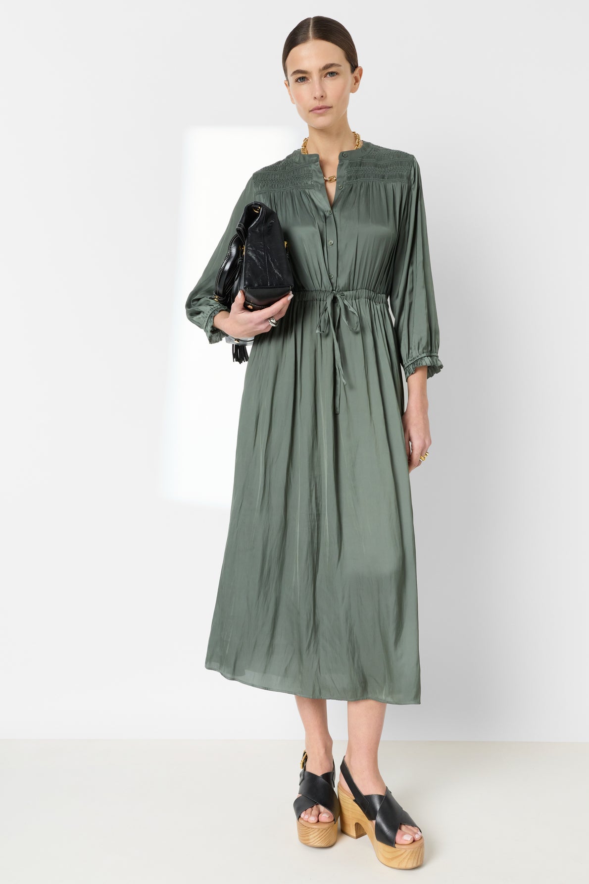 A person stands against a white wall wearing a robe satin kaki, a long-sleeved button-down dress with a cinched waist, black platform sandals, and holding a black handbag.
