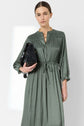 Woman wearing a long-sleeved, olive green robe satin kaki midi dress with a cinched waist, holding a black handbag, standing against a plain light gray background.