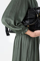 A person in a long-sleeved, olive green robe satin kaki dress holds a black leather handbag with white stitching and gold hardware, visible from the shoulders to mid-thigh against a plain background.