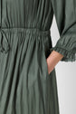 A close-up of a person wearing an olive green, long-sleeved robe longue with gathered fabric and a cinched waist, showing their hand in a side pocket.
