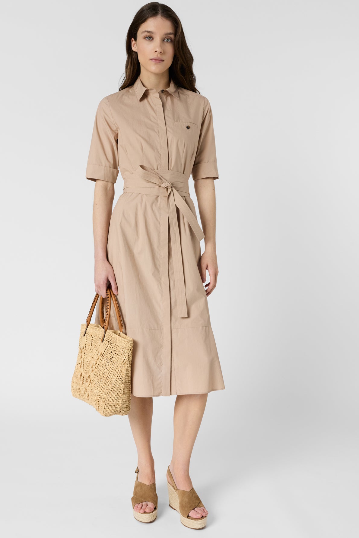 Woman wearing a beige collared midi dress with a waist tie, holding a woven handbag, and wearing tan wedge sandals, standing against a plain white background.