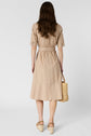 A woman stands facing away, wearing a beige knee-length dress with short sleeves and a belt. She holds a woven straw bag and wears wedge sandals. Her long, wavy brown hair is loose. The background is plain white.