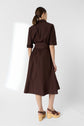 Woman standing with her back to the camera, wearing a dark brown, knee-length dress with rolled sleeves and a belt, plus brown platform sandals. Background is plain and white.