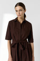 A woman with light skin and straight brown hair, tied back, wears a brown collared dress with short sleeves and a waist tie. She stands against a plain, light background, looking at the camera with a neutral expression.