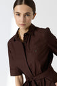 Woman with light eyes and straight brown hair pulled back, wearing a dark brown collared shirt dress with a pocket and gold chain necklace, standing against a plain light background.
