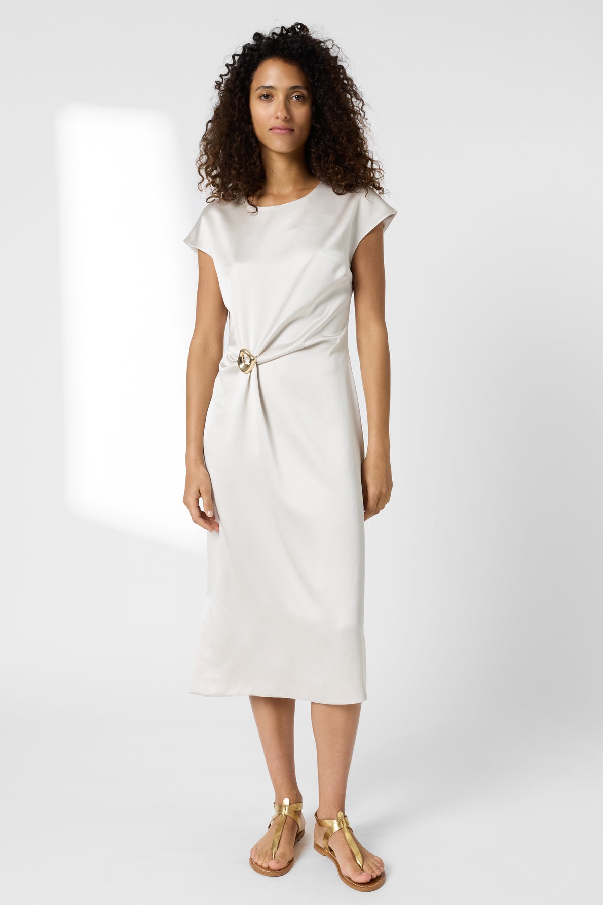 A woman with curly hair wears a light beige, knee-length satin dress with a knotted detail at the waist and gold sandals, standing against a plain white background.
